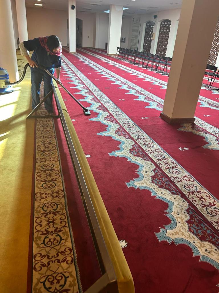 A person vacuuming a red carpet in a spacious indoor area, with rows of chairs in the background and intricate carpet designs.